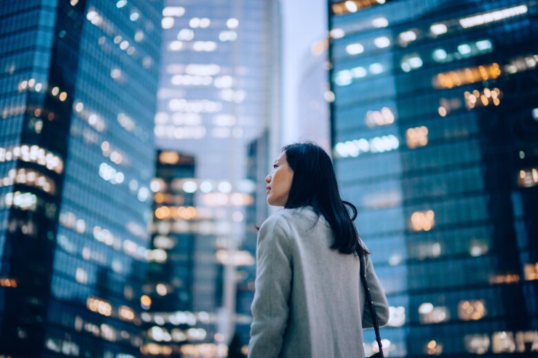 side profile of confident and professional young Asian businesswoman looking up while standing against contemporary corporate skyscrapers