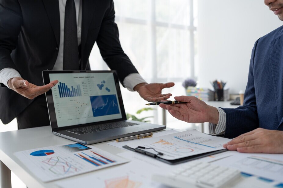 Two people reviewing business performance data during an office meeting. A laptop displays charts and analytics dashboards while printed reports with graphs and metrics are spread across a conference table. One person gestures toward the data as another holds a pen while discussing results.