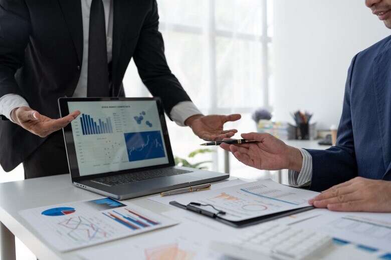 Two people reviewing business performance data during an office meeting. A laptop displays charts and analytics dashboards while printed reports with graphs and metrics are spread across a conference table. One person gestures toward the data as another holds a pen while discussing results.