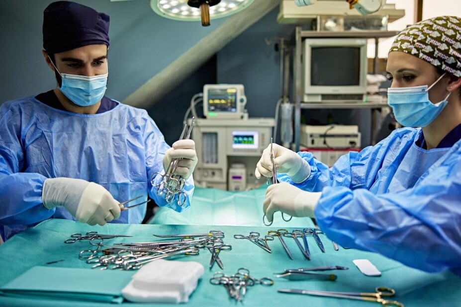 Waist up view of young doctors in masks, caps, gloves, and operating gowns arranging tray of scalpels, scissors, forceps, and clamps in operating room.
