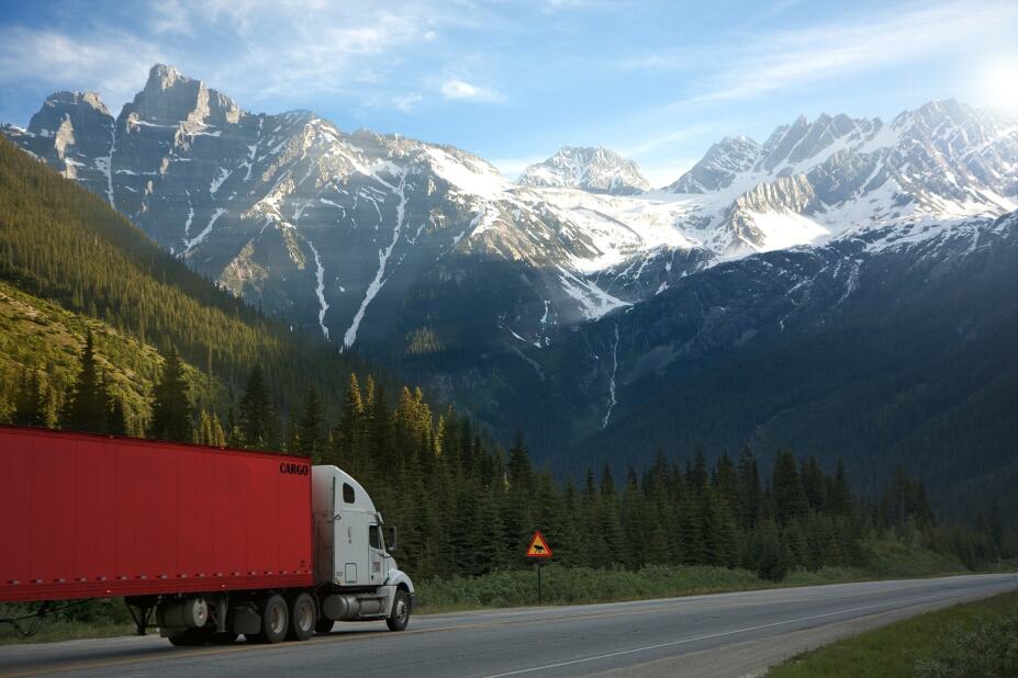 tractor trailer truck driving down a highway with forest and snow-capped mountains in the background