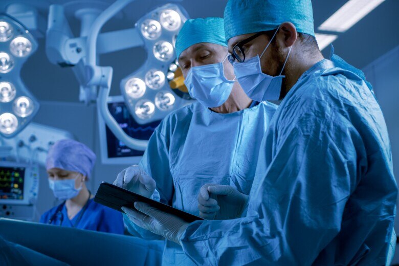 Professional Surgeons and Assistants Talk and Use Digital Tablet Computer while Standing in the Modern Hospital Operating Room.