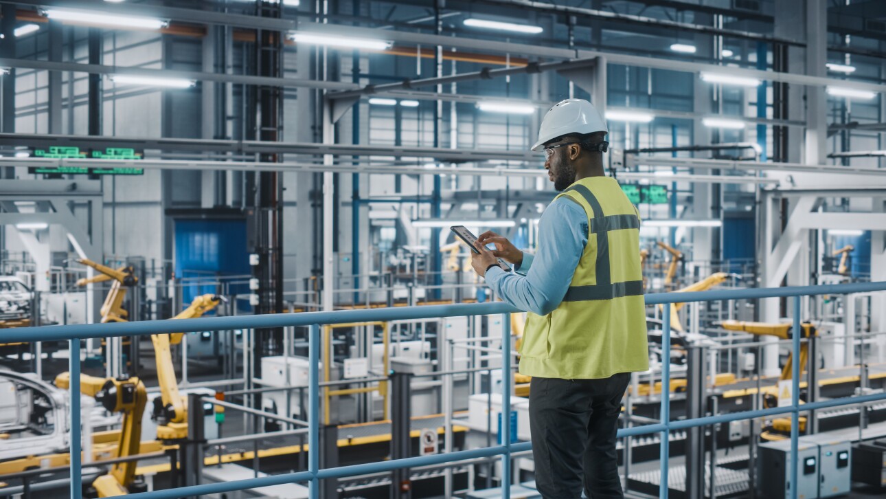 African American Car Factory Engineer in High Visibility Vest Using Tablet Computer. Automotive Industrial Manufacturing Facility Working on Vehicle Production on Automated Technology Assembly Plant.